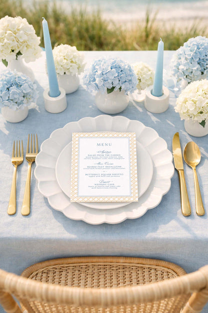 a beautifully set table for a formal dining experience. The table is adorned with a blue and white color scheme, and the centerpiece is a vase filled with blue hydrangeas. The table is set with a white plate, gold cutlery, and a menu card. The overall scene conveys a sense of elegance and sophistication.