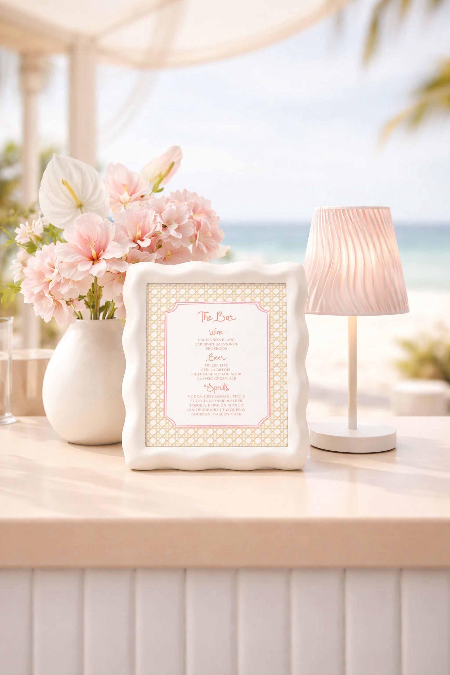 a white table with a framed menu, a vase of pink flowers, and a lamp, all placed on a beach table.