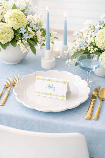A table setting with a white plate, goldware, and a card with the name "Harley" written on it. The table is decorated with white flowers, blue candles, and a blue vase.