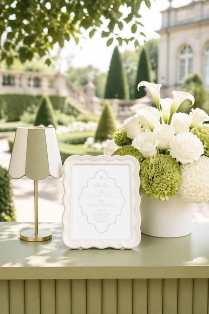 a white frame with a floral arrangement of white roses and green hydrangeas, placed on a table next to a lamp. The background includes a well-manicured garden with trees and a building, creating a serene and elegant setting.
