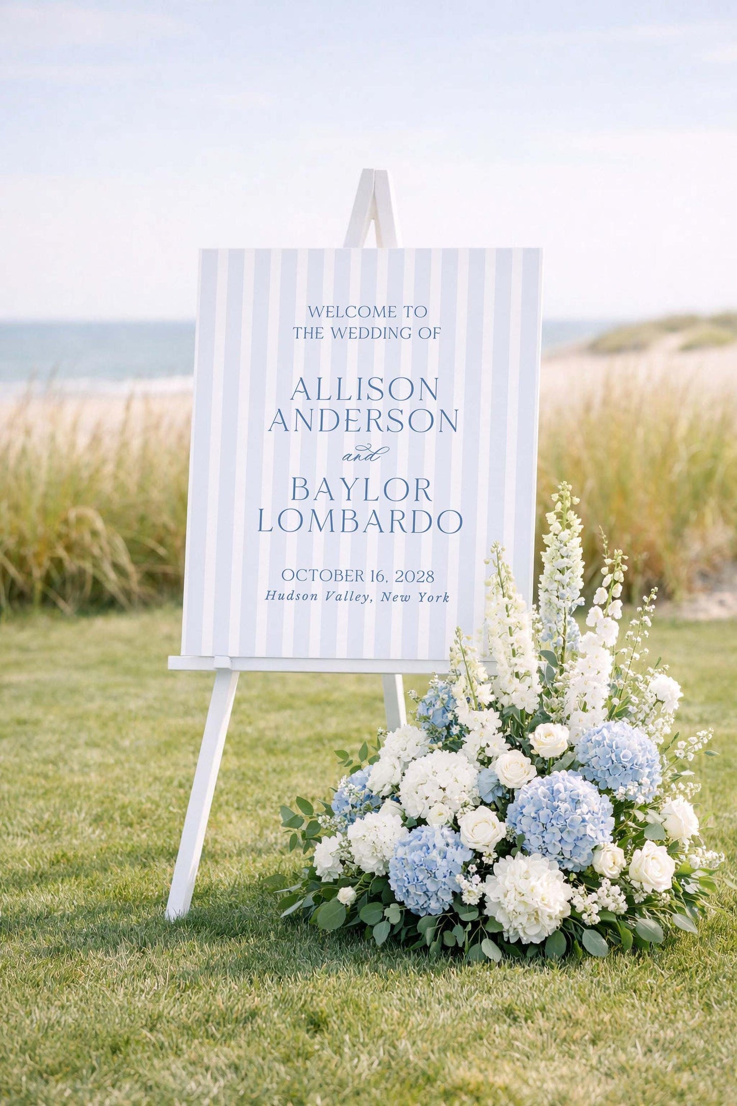 a white sign with blue stripes welcoming guests to the wedding of Allison Anderson and Baylor Lombardo. The sign is set against a backdrop of a grassy field and a beach, with a beautiful flower arrangement in the foreground.
