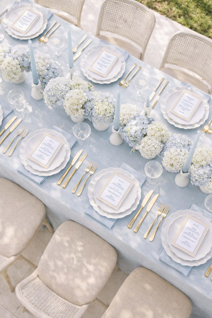 a beautifully set dining table with white plates, gold cutlery, and a floral centerpiece. The table is surrounded by beige chairs, and the table is set for a formal event or celebration.
