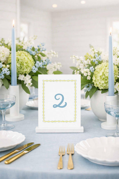 a table setting with a white tablecloth, a white table number holder displaying the number 2, and various tableware items such as plates, glasses, and silverware.