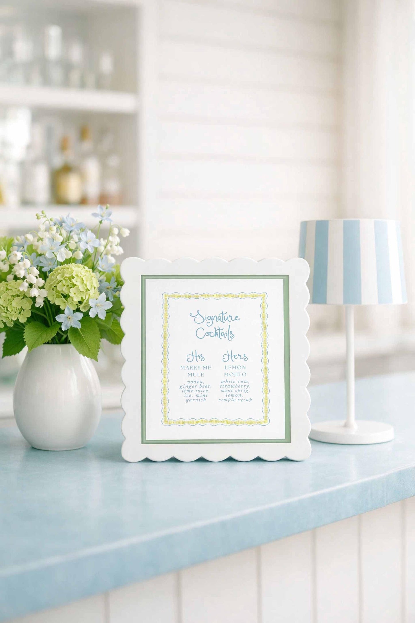 a white scalloped frame with a list of cocktail names, placed on a blue countertop next to a vase of white flowers and a white lamp.