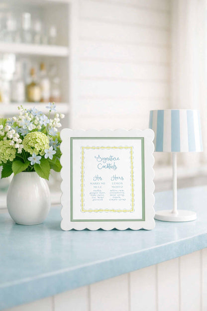 a white scalloped frame with a list of cocktail names, placed on a blue countertop next to a vase of white flowers and a white lamp.