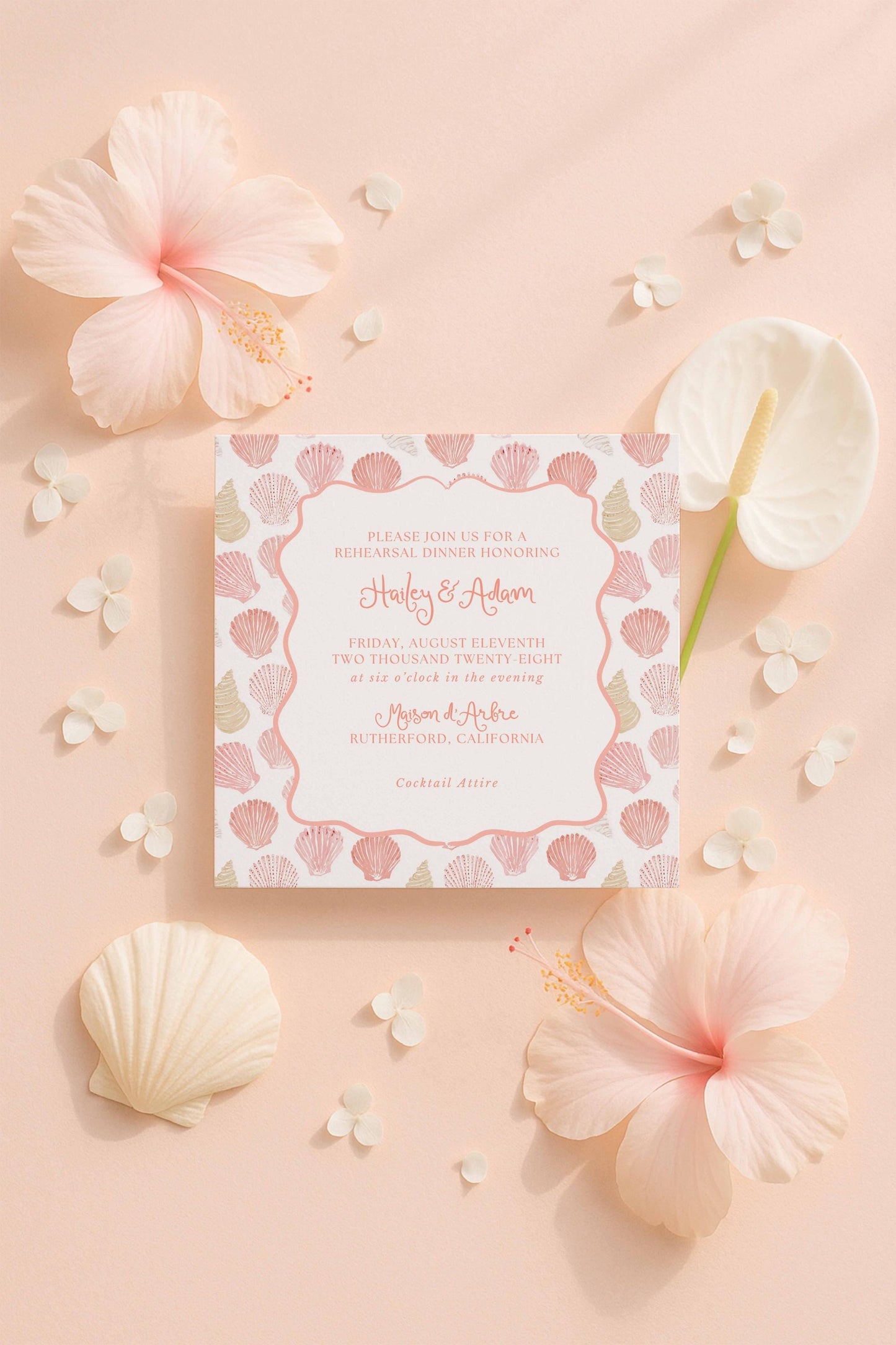a wedding invitation card with a pink and white floral design, surrounded by various pink and white flowers, shells, and a green pen.
