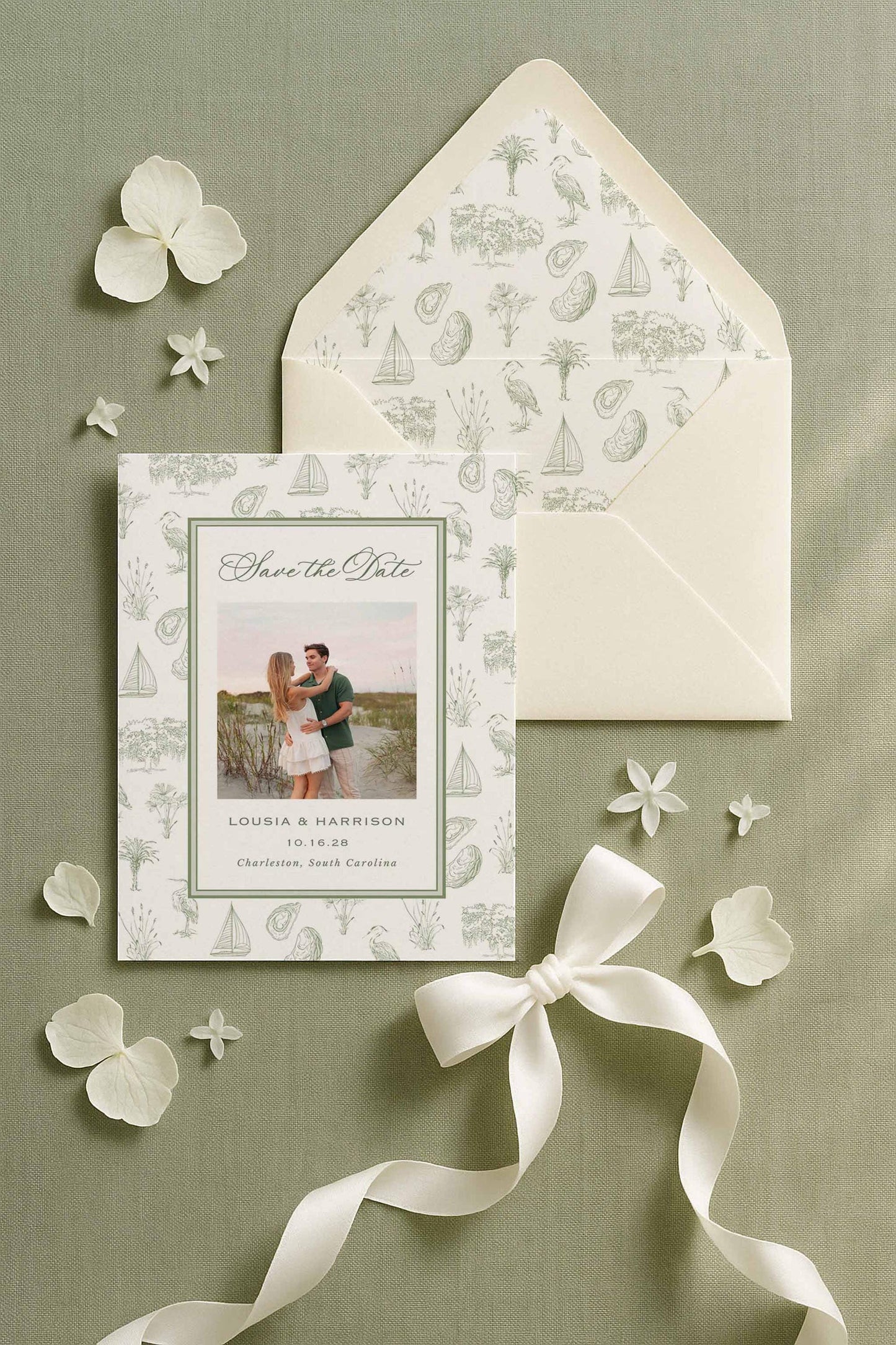 Southern toile Save the Date card displayed on a sage green linen background with white hydrangea petals and satin ribbon. The design features a fine art toile border of hand-drawn palmettos, egrets, oysters, oak trees, sailboats, and marsh grasses. Shown in a no-photo layout with wedding date and names centered. Printable and Canva-editable template ideal for southern weddings, coastal events, and Lowcountry-inspired stationery suites. Envelope liner included for a matching look.