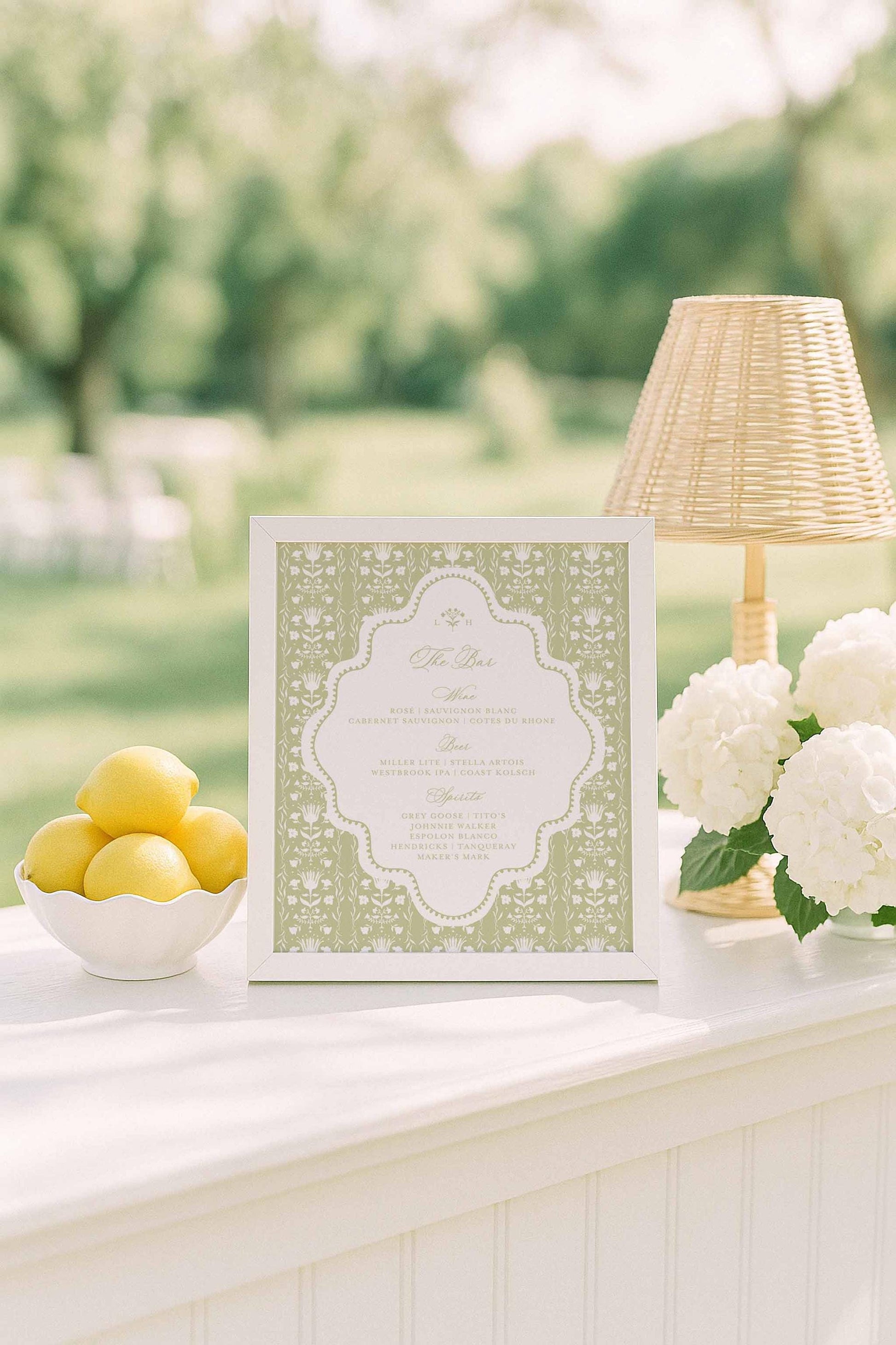a white table with a framed green and white invitation, a bowl of lemons, and a lamp, all set against a blurred background of trees and greenery.