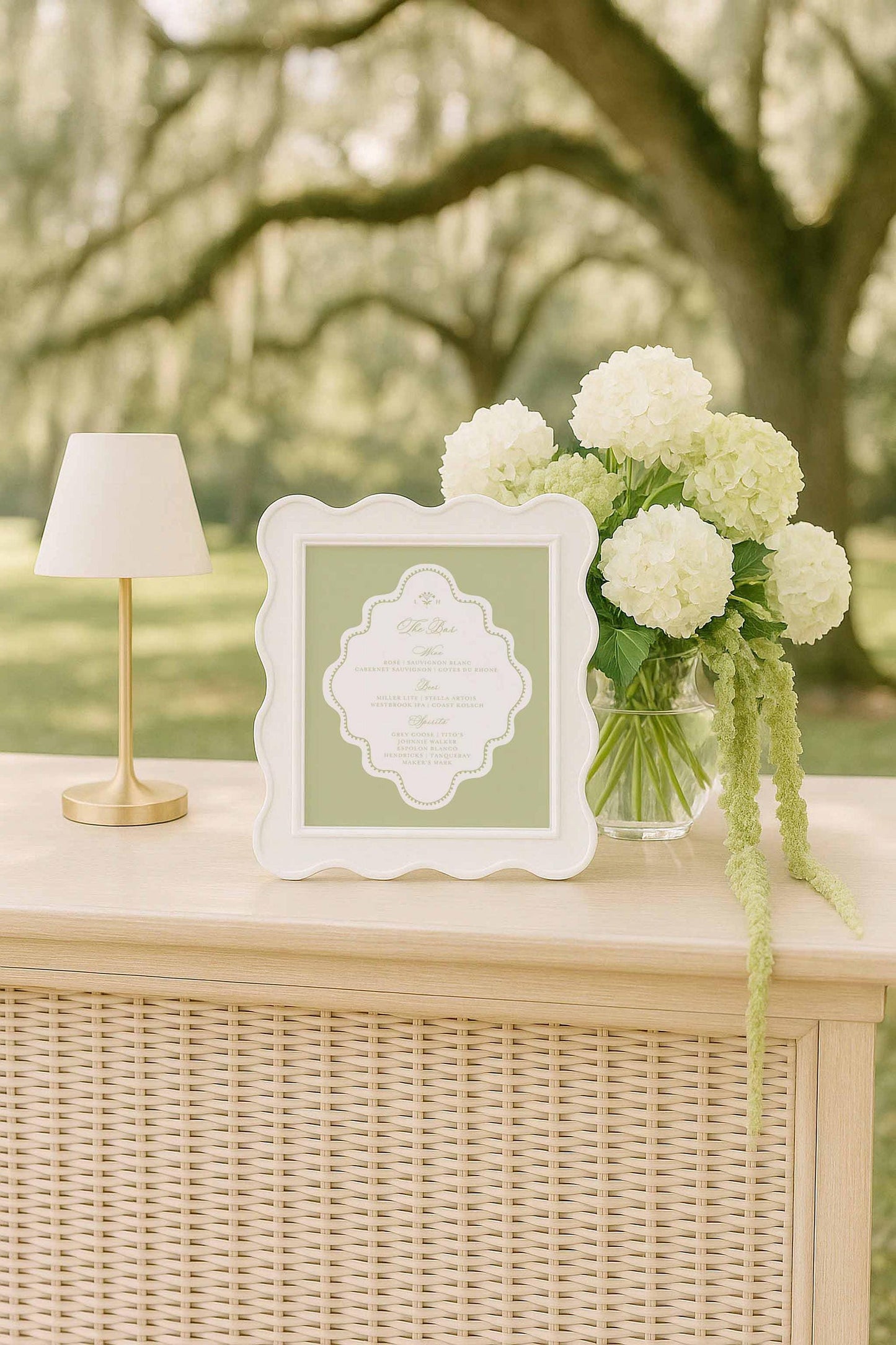 a white frame with a green border, placed on a wooden table, accompanied by a vase of white hydrangeas and a lamp.