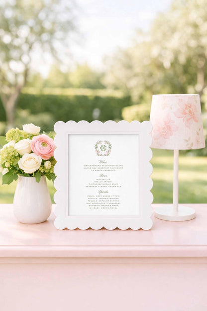 a white frame with a floral design, a vase of flowers, and a lamp on a pink table.