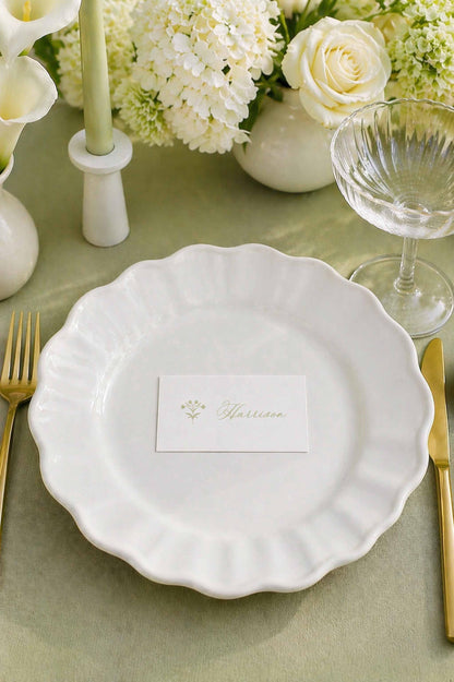 a beautifully set table with a white plate, a name card, a glass of wine, and a vase of white flowers.