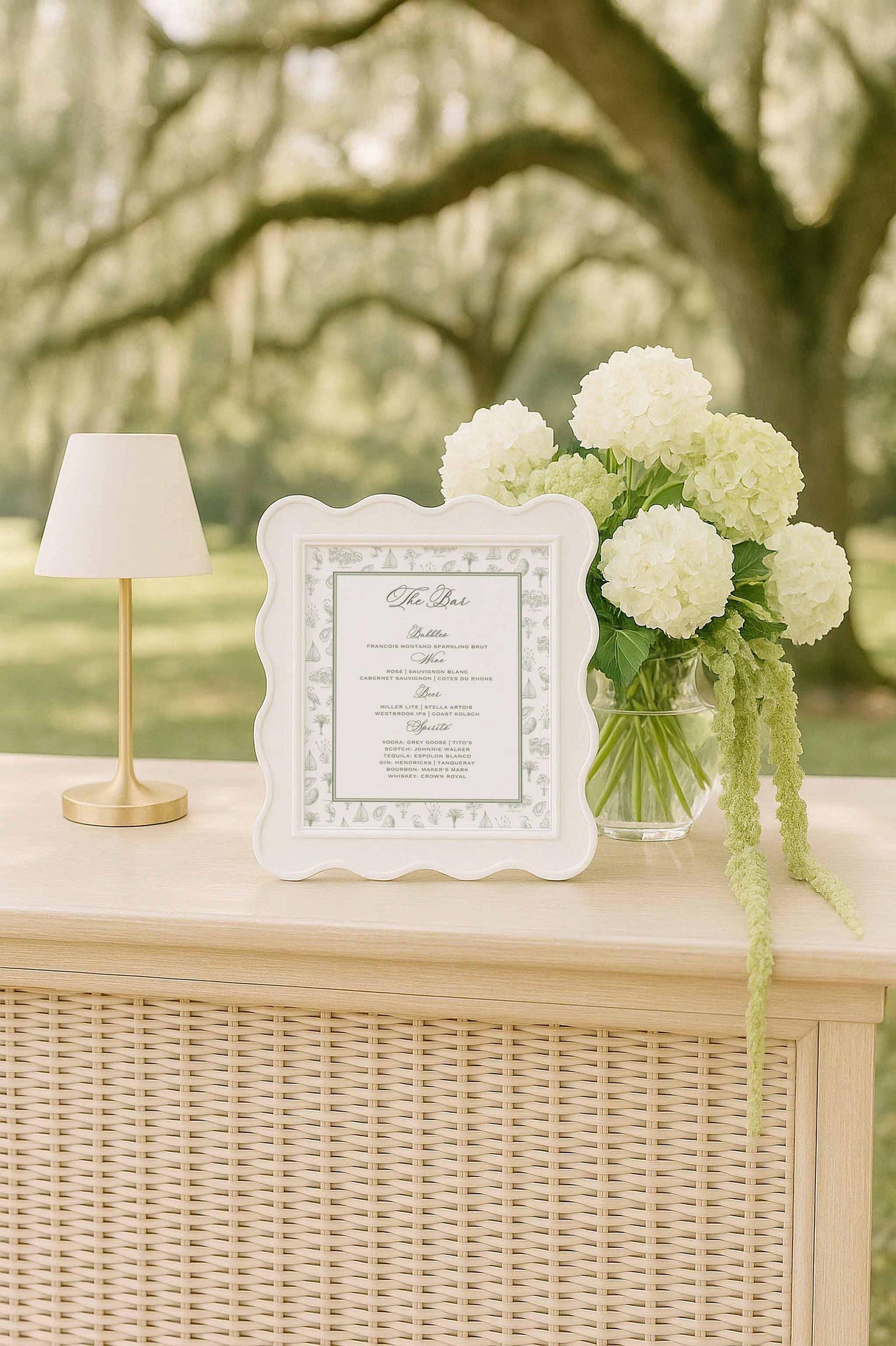 Wedding bar menu with southern toile artwork featuring palmettos, egrets, oysters, oak trees, marsh scenes, and sailboats. Displayed in a white scalloped frame on a rattan bar with soft green and white florals and a striped lamp. Fine art coastal stationery with customizable drink selections. Printable and Canva-editable bar menu sign ideal for southern weddings, outdoor receptions, and elegant coastal celebrations.
