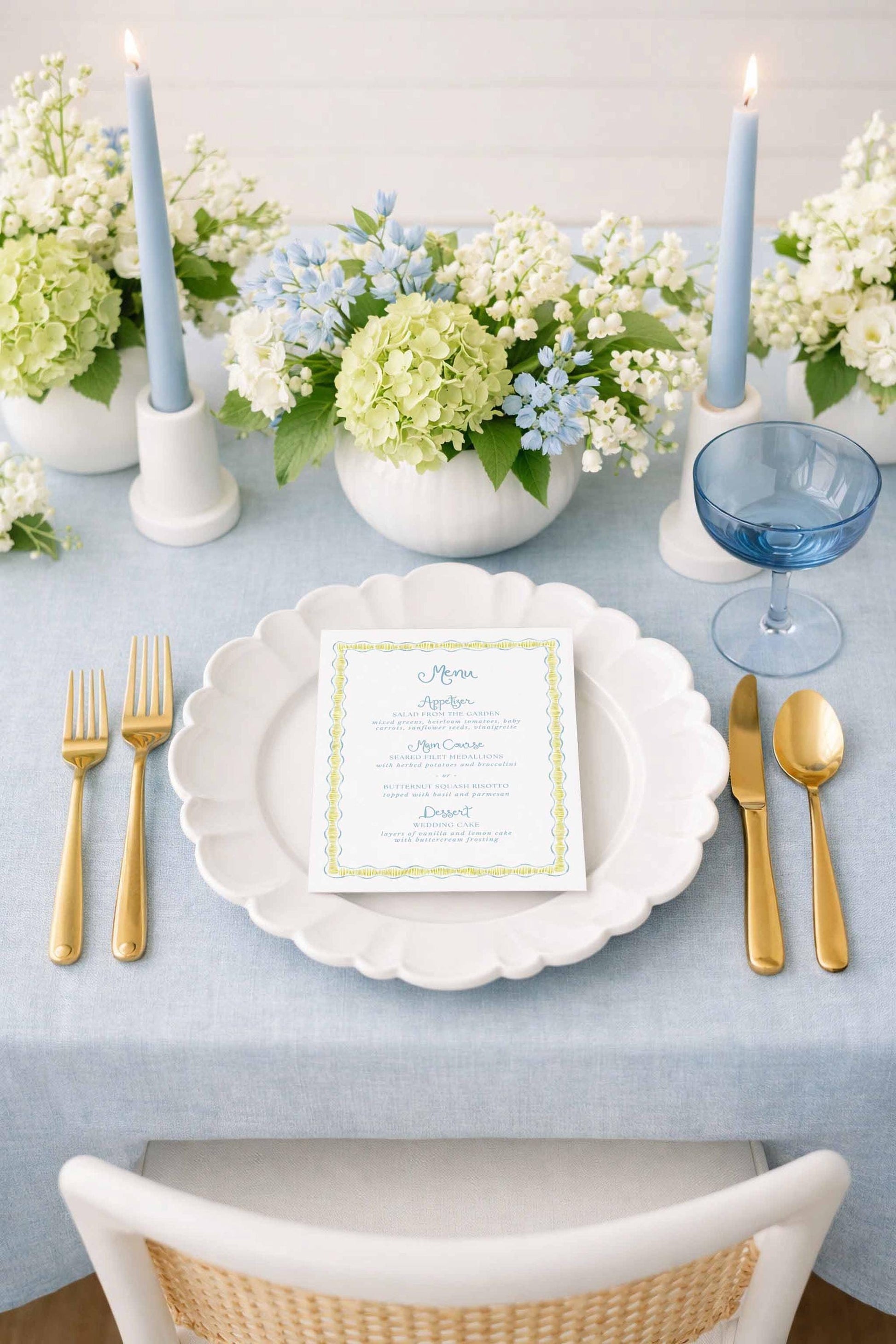 a beautifully set dining table with a white plate, gold cutlery, and a menu card. The table is adorned with blue and white flowers, and there are two blue candles on either side of the plate. The overall setting appears elegant and inviting.