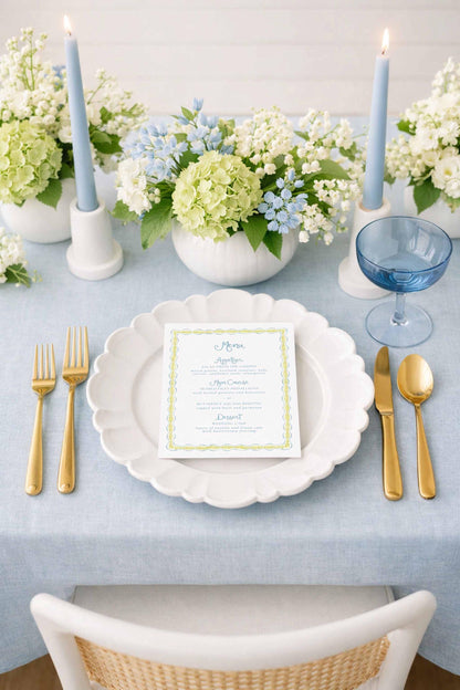 a beautifully set dining table with a white plate, gold cutlery, and a menu card. The table is adorned with blue and white flowers, and there are two blue candles on either side of the plate. The overall setting appears elegant and inviting.