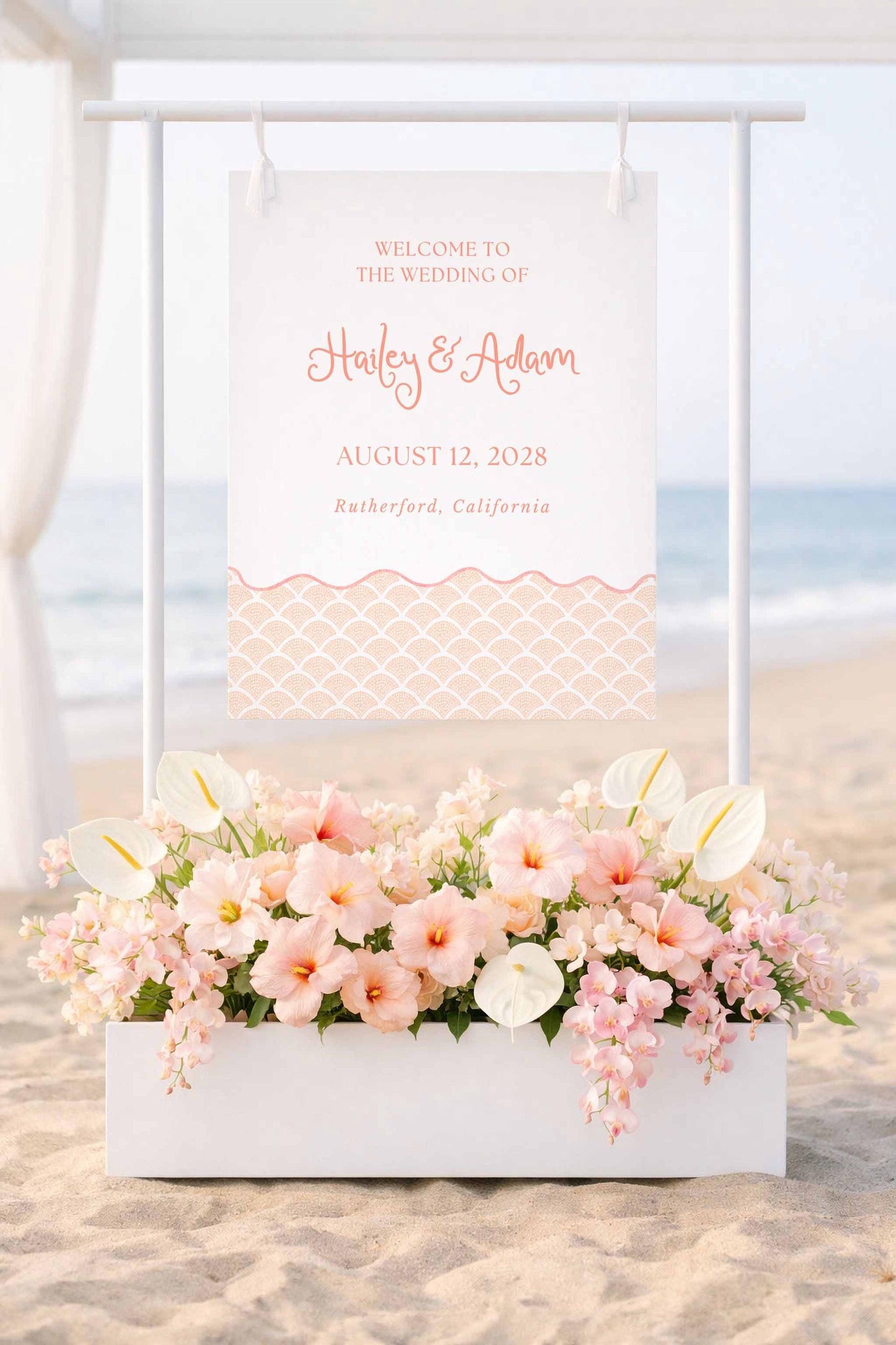 a wedding invitation sign with a beach scene and a white planter filled with pink and white flowers in the foreground.