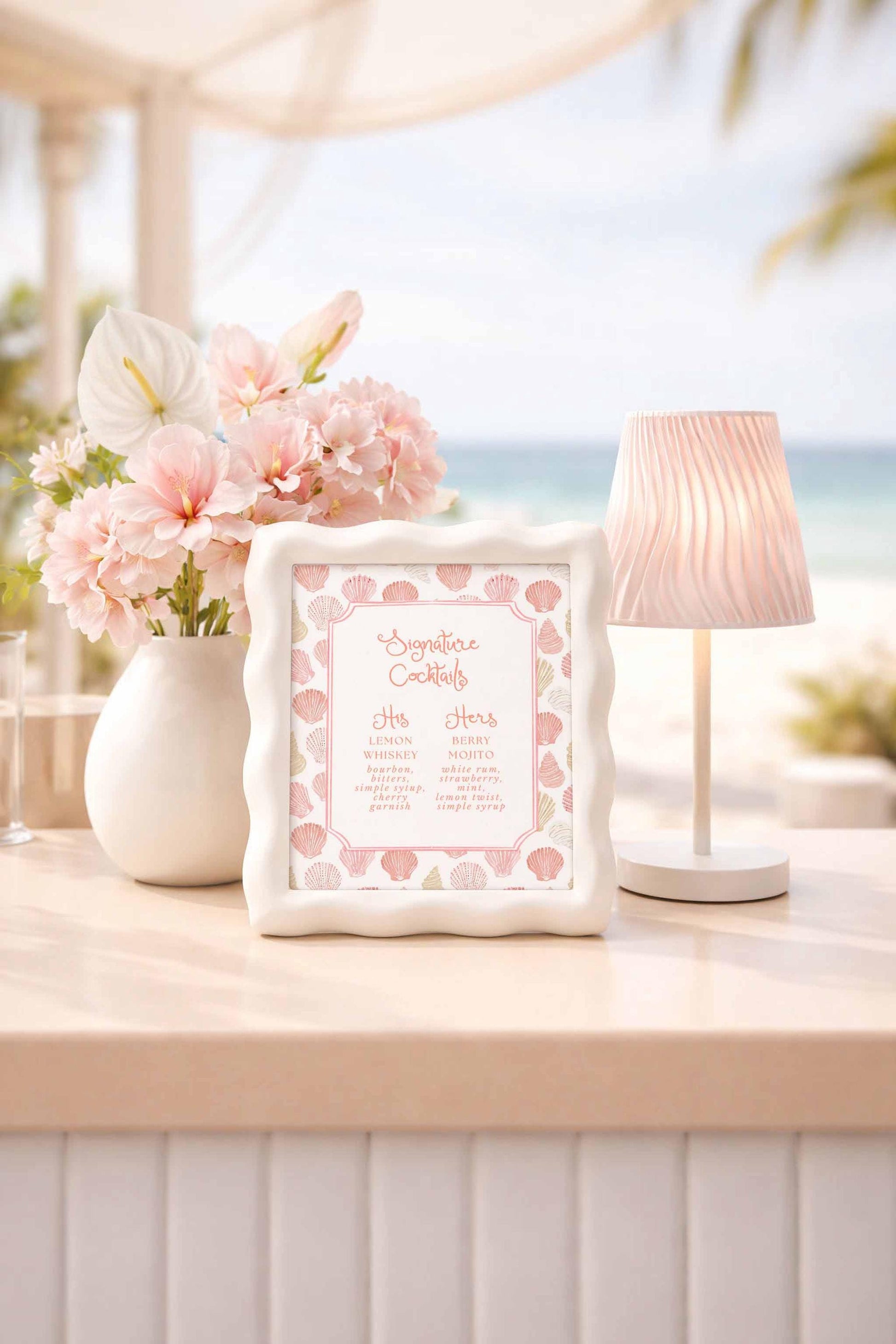 a table with a framed cocktail menu, a vase of pink flowers, and a lamp, all set against a beach background.