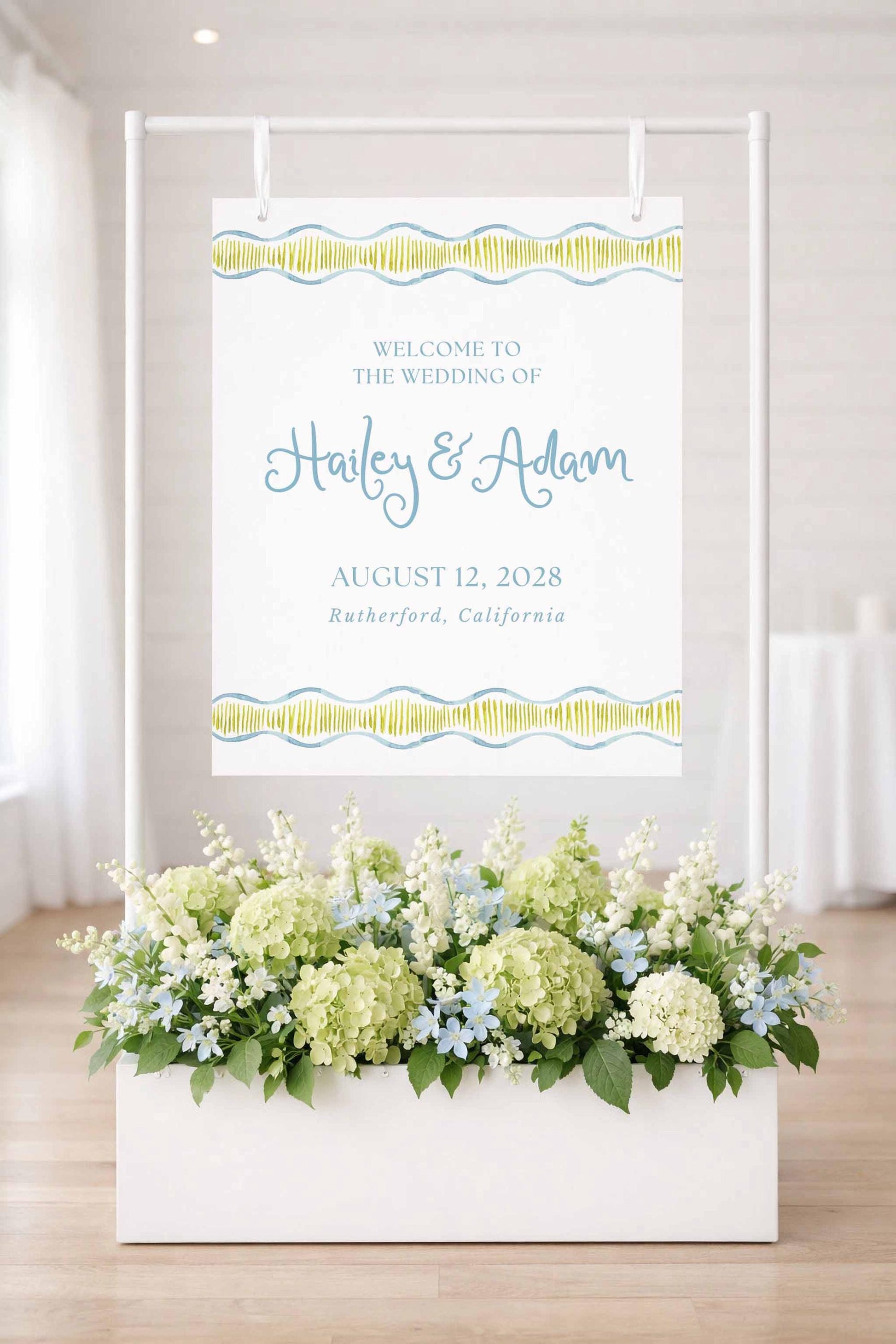 a white sign with a floral arrangement in a white planter, displaying the text "Welcome to the wedding of Haley & Adam" and the date "August 12, 2028" in a decorative font.