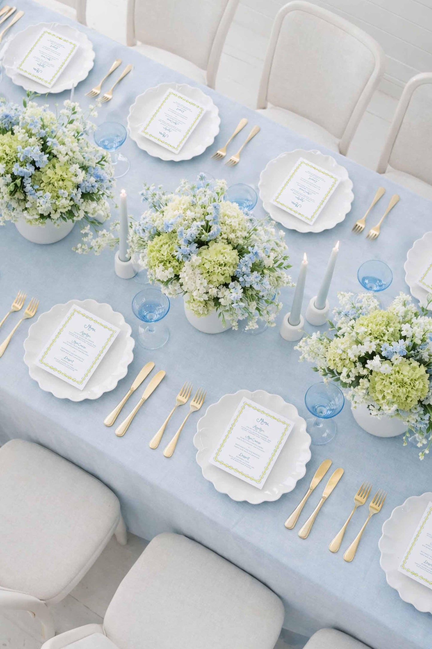 a beautifully set dining table with a light blue tablecloth, white plates, and gold cutlery. The table is adorned with white flowers in blue and white vases, and there are blue and white place cards and glasses arranged on the table.