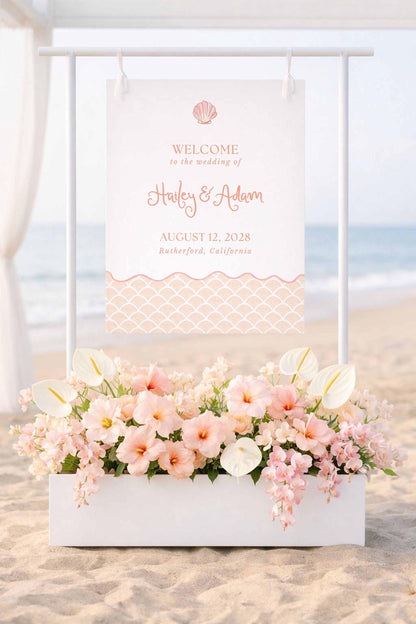 a wedding invitation sign on a beach, with a white signboard displaying the names "Harley & Adam" and the date "AUGUST 12, 2028" in a coastal theme. Below the signboard, there is a white planter filled with pink and white flowers, creating a beautiful and romantic atmosphere for the wedding ceremony.