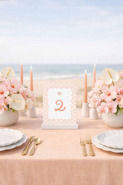 a table set for a formal dining experience, with a tablecloth, plates, silverware, and a centerpiece of pink flowers. The table is positioned near the ocean, creating a serene and picturesque setting.