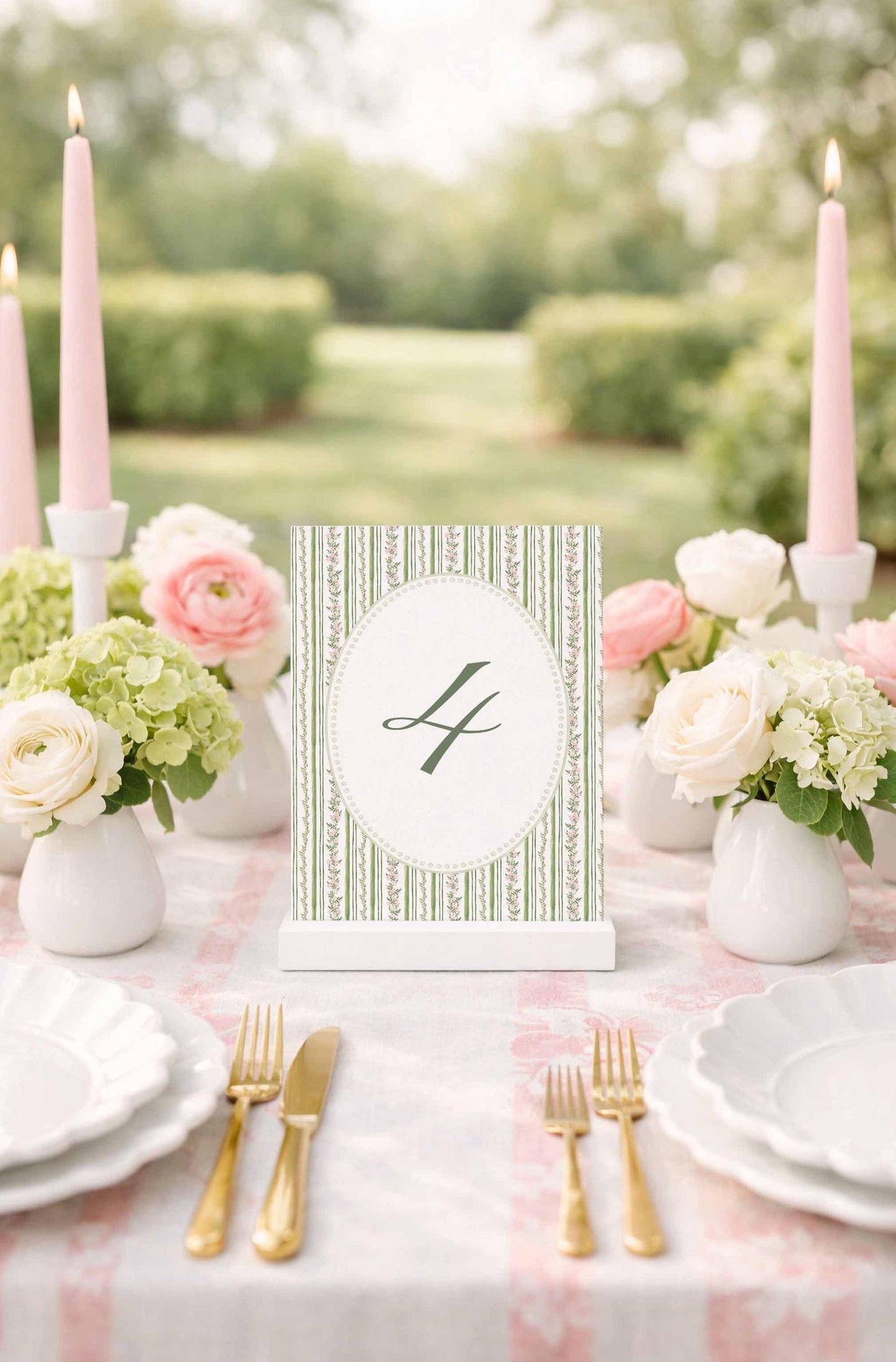 A table setting with a white tablecloth, pink candles, white plates, and pink and white flowers. A green and white striped table number holder is placed in the center of the table.