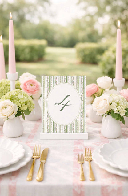 A table setting with a white tablecloth, pink candles, white plates, and pink and white flowers. A green and white striped table number holder is placed in the center of the table.