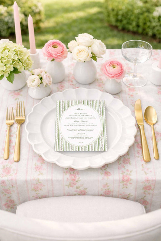 a beautifully set table with a white tablecloth, gold-colored cutlery, and a menu card. The table is adorned with pink and white flowers, and there are candles and a wine glass on the table as well.