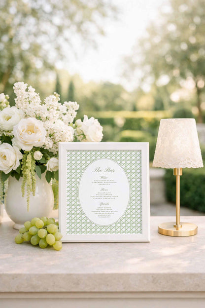 a white frame with a green pattern, a bouquet of white flowers, a bunch of green grapes, and a gold-colored lamp on a table.