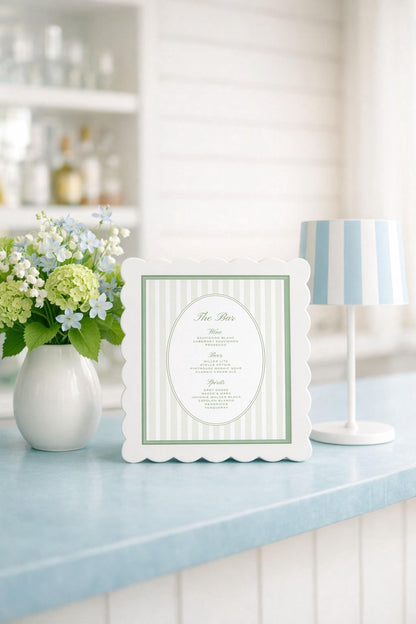 a white framed menu with a green border, placed on a blue countertop next to a vase of white and green flowers.
