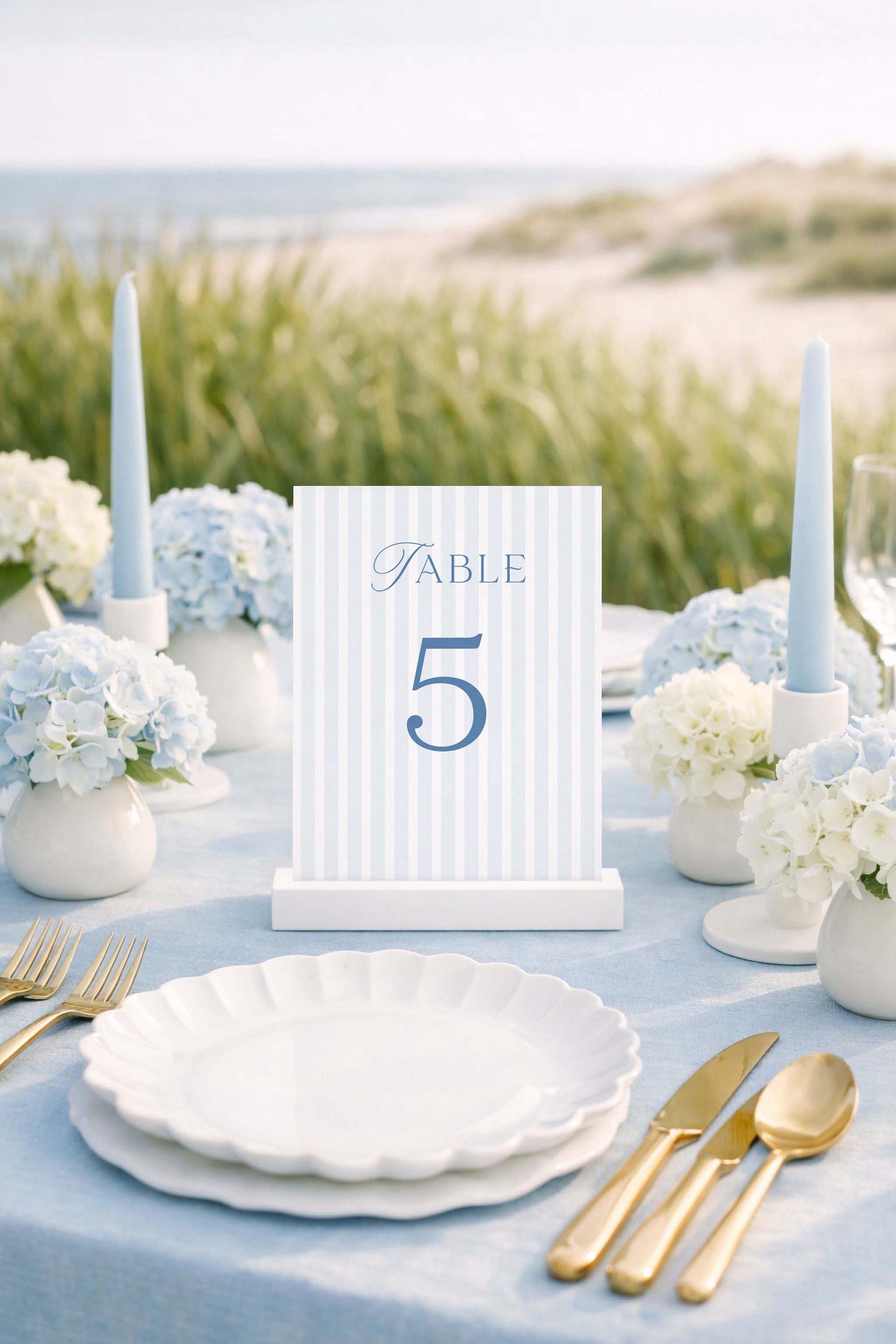 A table setting with a blue and white striped table number 5, white plates, gold cutlery, and white flowers, with a beach view in the background.