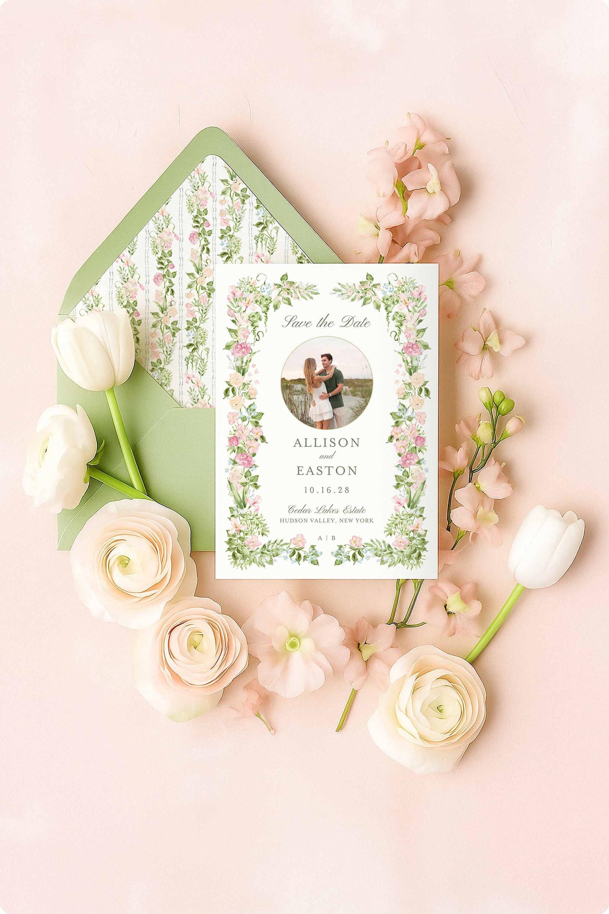A photo Save the Date card in a watercolor floral frame placed over a matching floral backer, surrounded by delicate blush flowers and white tulips. Elegant matching envelope liner included with green envelope.