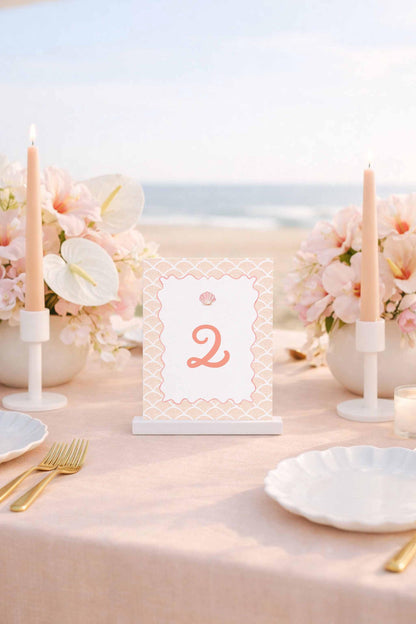 a table set for a romantic beachside dinner, with a pink tablecloth, white plates, and candles. A table number sign displaying the number '2' is placed in the center of the table.