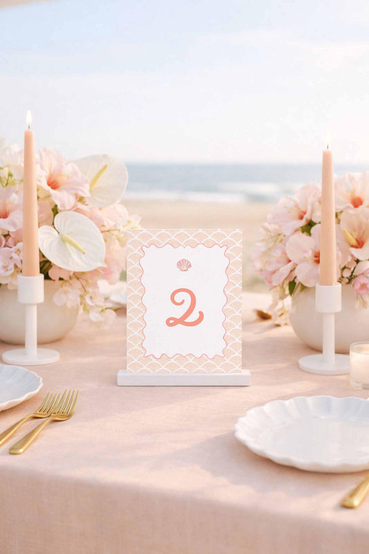 a table set for a romantic beachside dinner, with a pink tablecloth, white plates, and candles. A table number sign displaying the number '2' is placed in the center of the table.