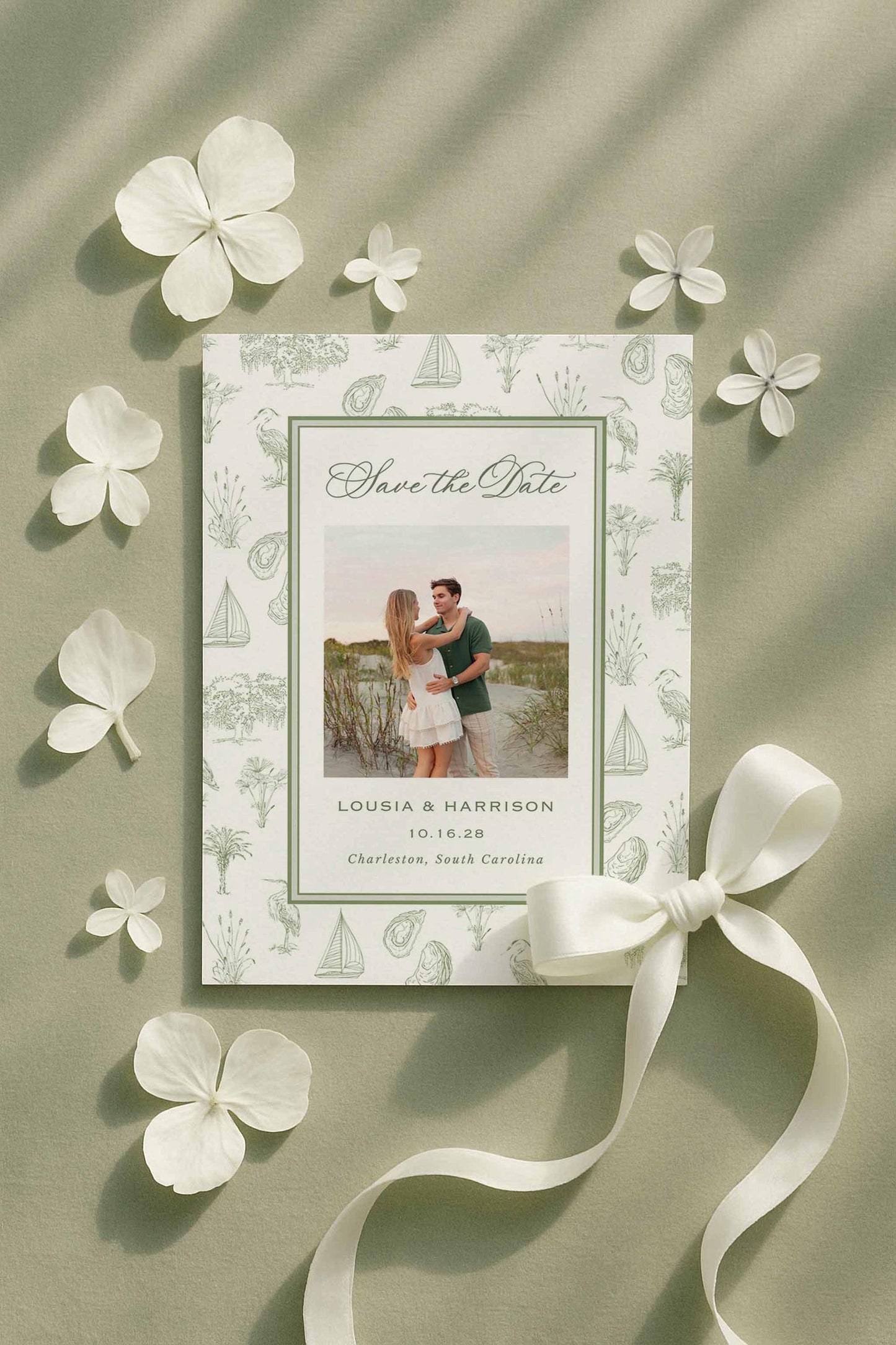 Save the Date card with southern toile artwork surrounding a customizable text area and optional engagement photo. Displayed alongside white flower petals and a satin ribbon on green fabric. The toile design includes palmettos, egrets, oysters, sailboats, oak trees, and marsh elements. Canva editable, printable, and includes a matching toile envelope liner for a cohesive wedding stationery suite. Perfect for coastal and Lowcountry weddings.
