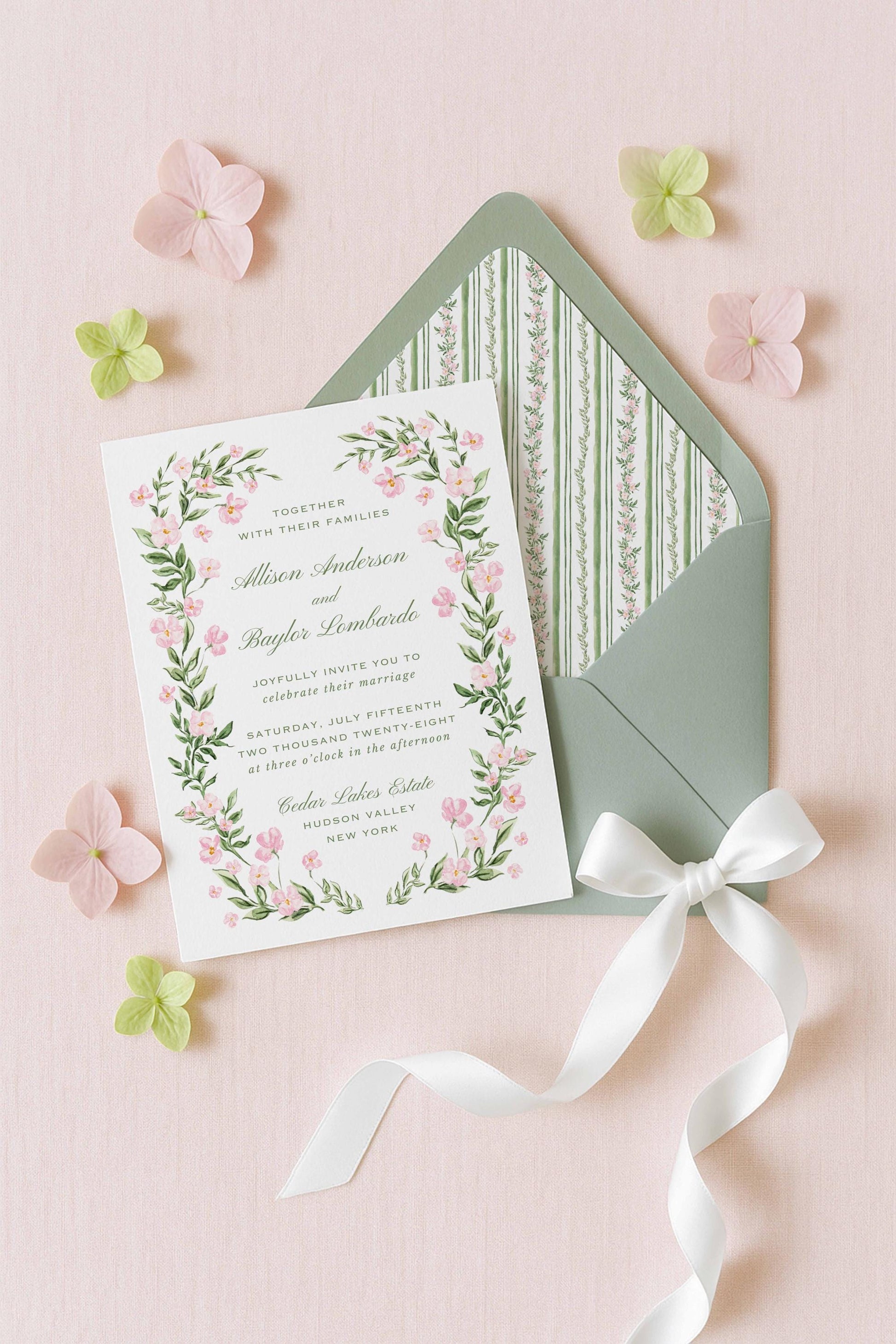 A white wedding invitation card with a floral border and a green envelope with a white ribbon bow, surrounded by pink and green flowers on a pink background.