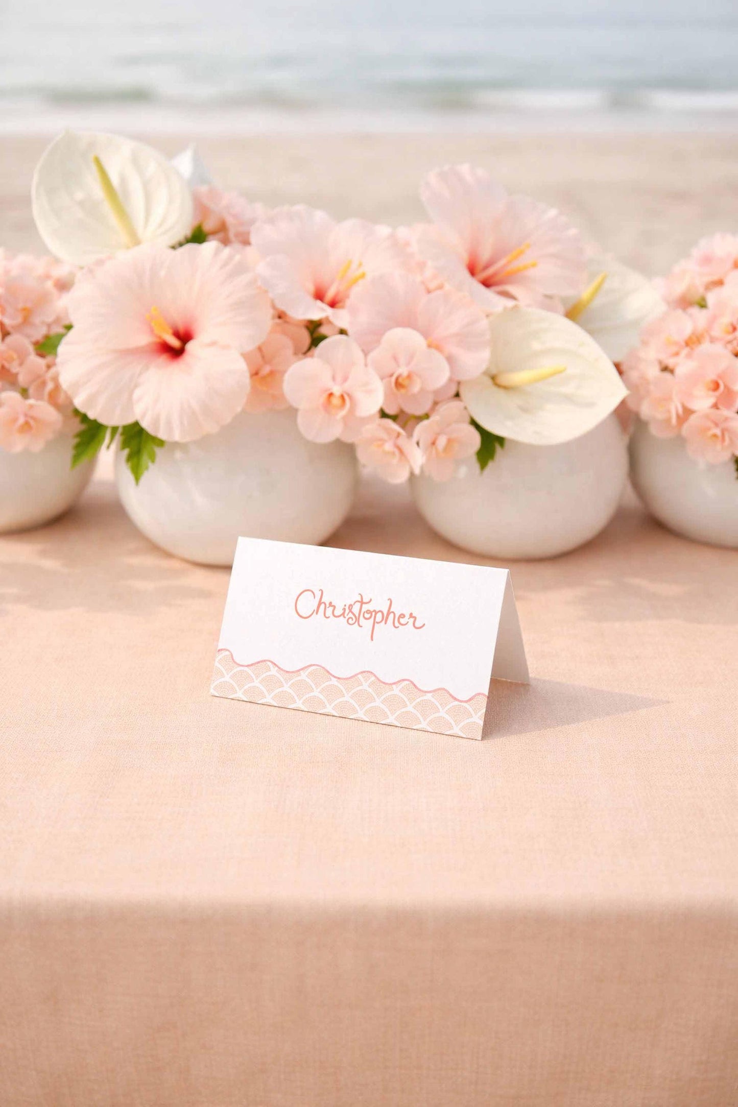 a table with a white tablecloth and a small white card with the name "Christopher" written on it. In the background, there are several vases filled with pink flowers, creating a beautiful and elegant setting.