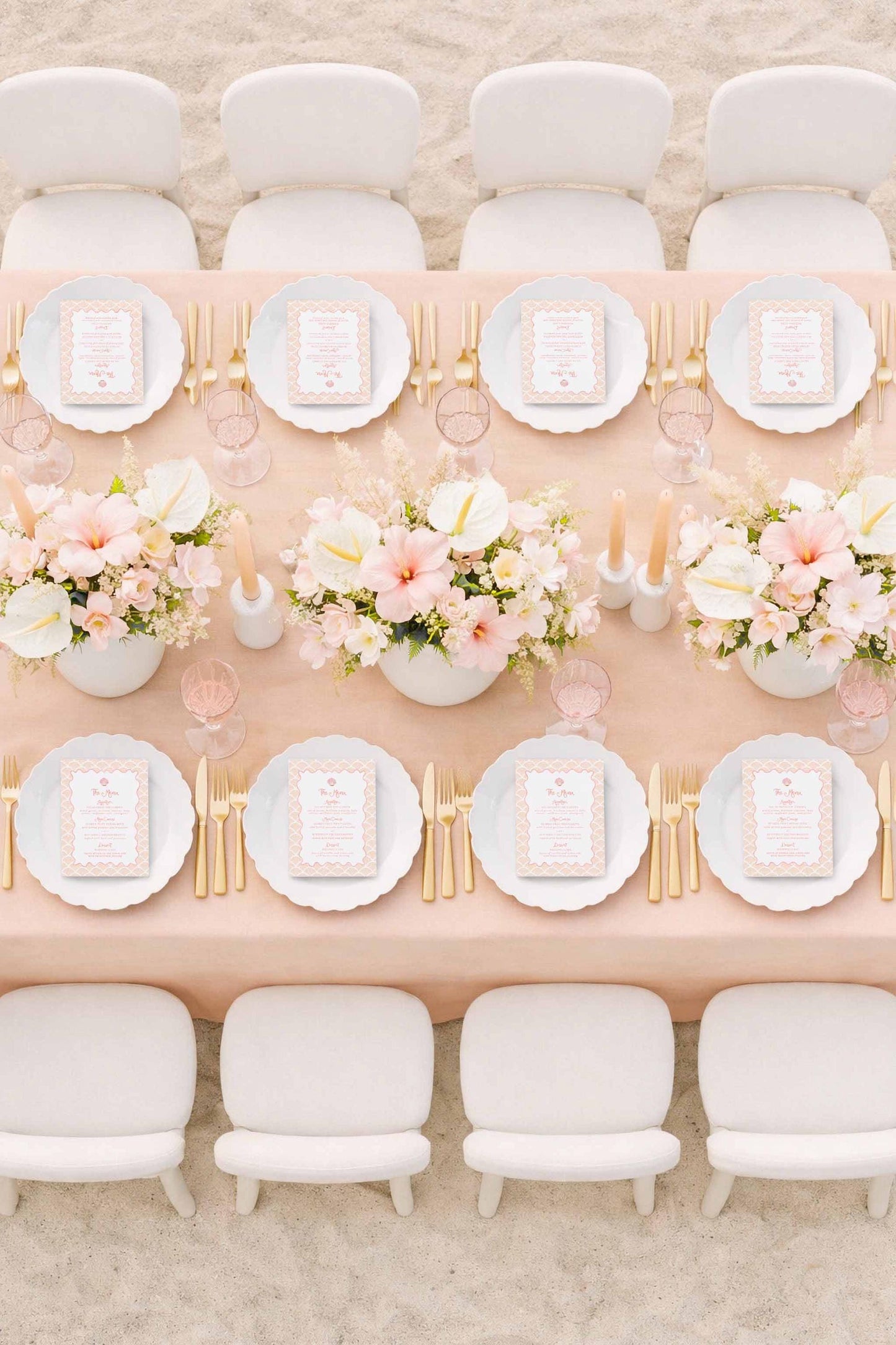 a beautifully set dining table with white plates, gold cutlery, and a vase of pink and white flowers. The table is surrounded by white chairs, creating an elegant and inviting atmosphere.