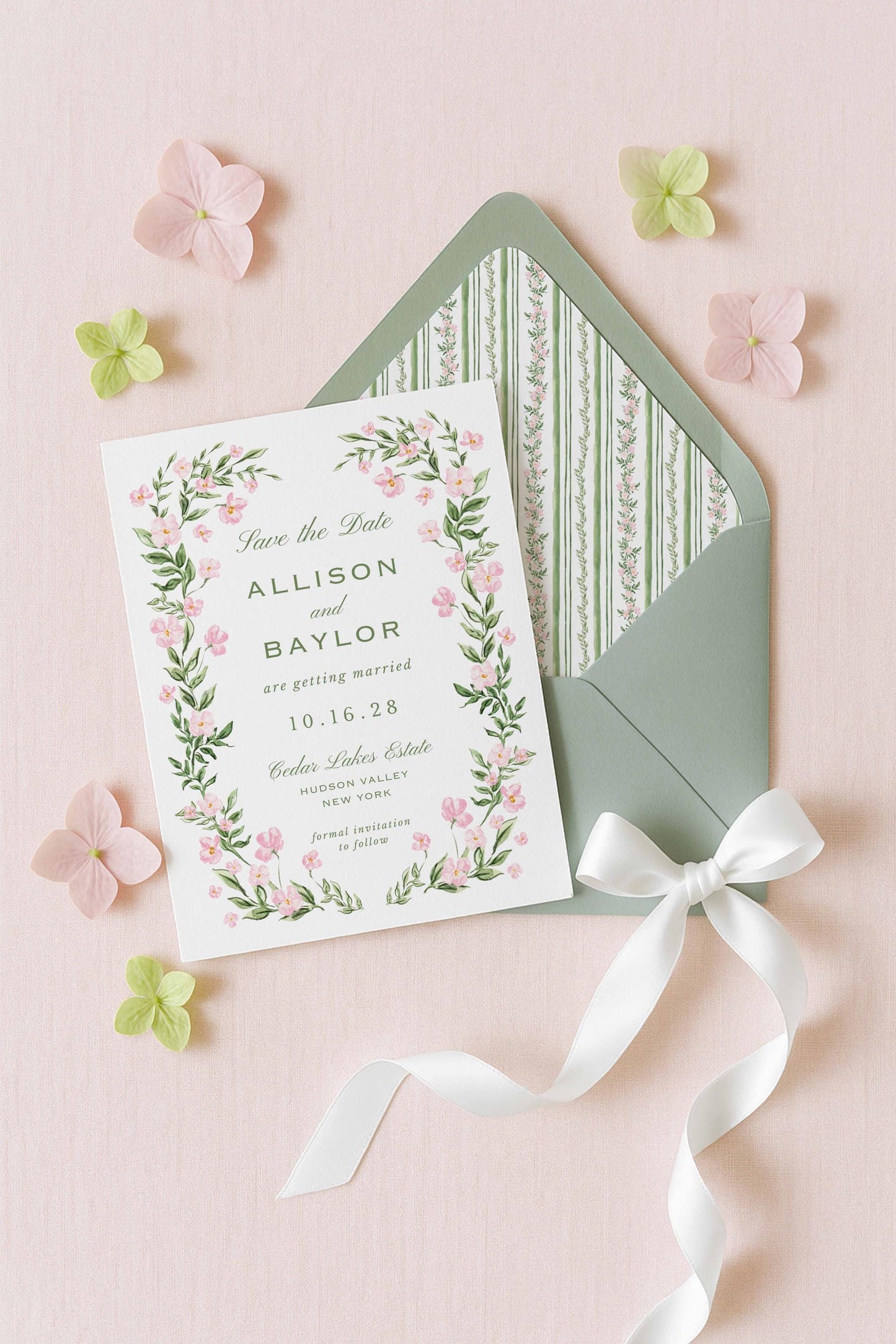 A white wedding invitation card with a floral border and a green envelope with a white ribbon, surrounded by pink and green flowers on a pink background.