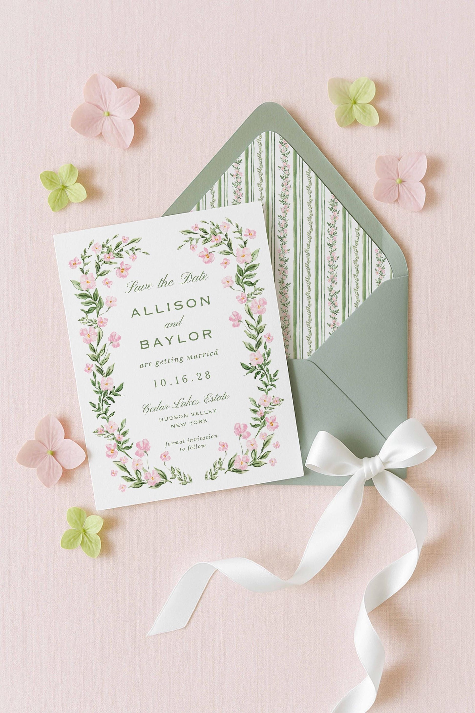 A white wedding invitation card with a floral border and a green envelope with a white ribbon, surrounded by pink and green flowers on a pink background.