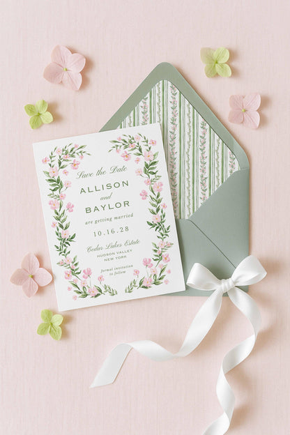A white wedding invitation card with a floral border and a green envelope with a white ribbon, surrounded by pink and green flowers on a pink background.
