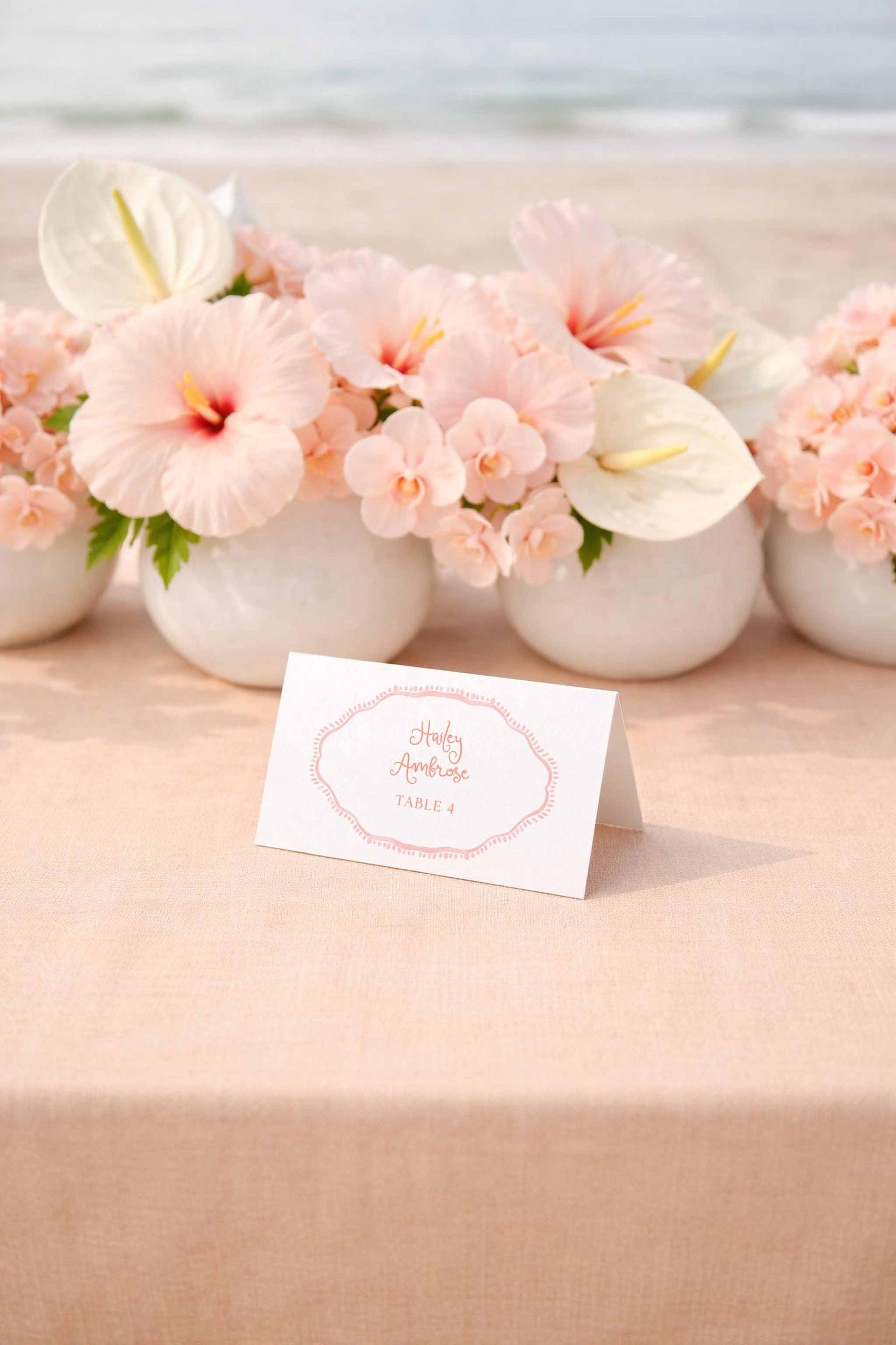 a table with a white card in the foreground displaying the text "Holly & Andy's Table 4" surrounded by a floral arrangement of pink hibiscus flowers and white orchids.