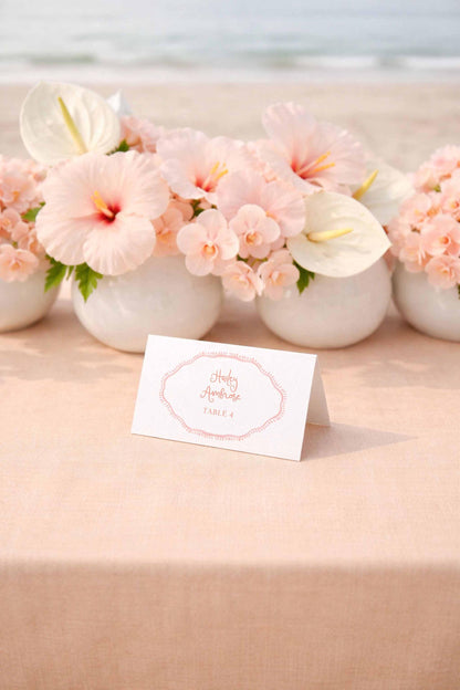 a table with a white card in the foreground displaying the text "Holly & Andy's Table 4" surrounded by a floral arrangement of pink hibiscus flowers and white orchids.