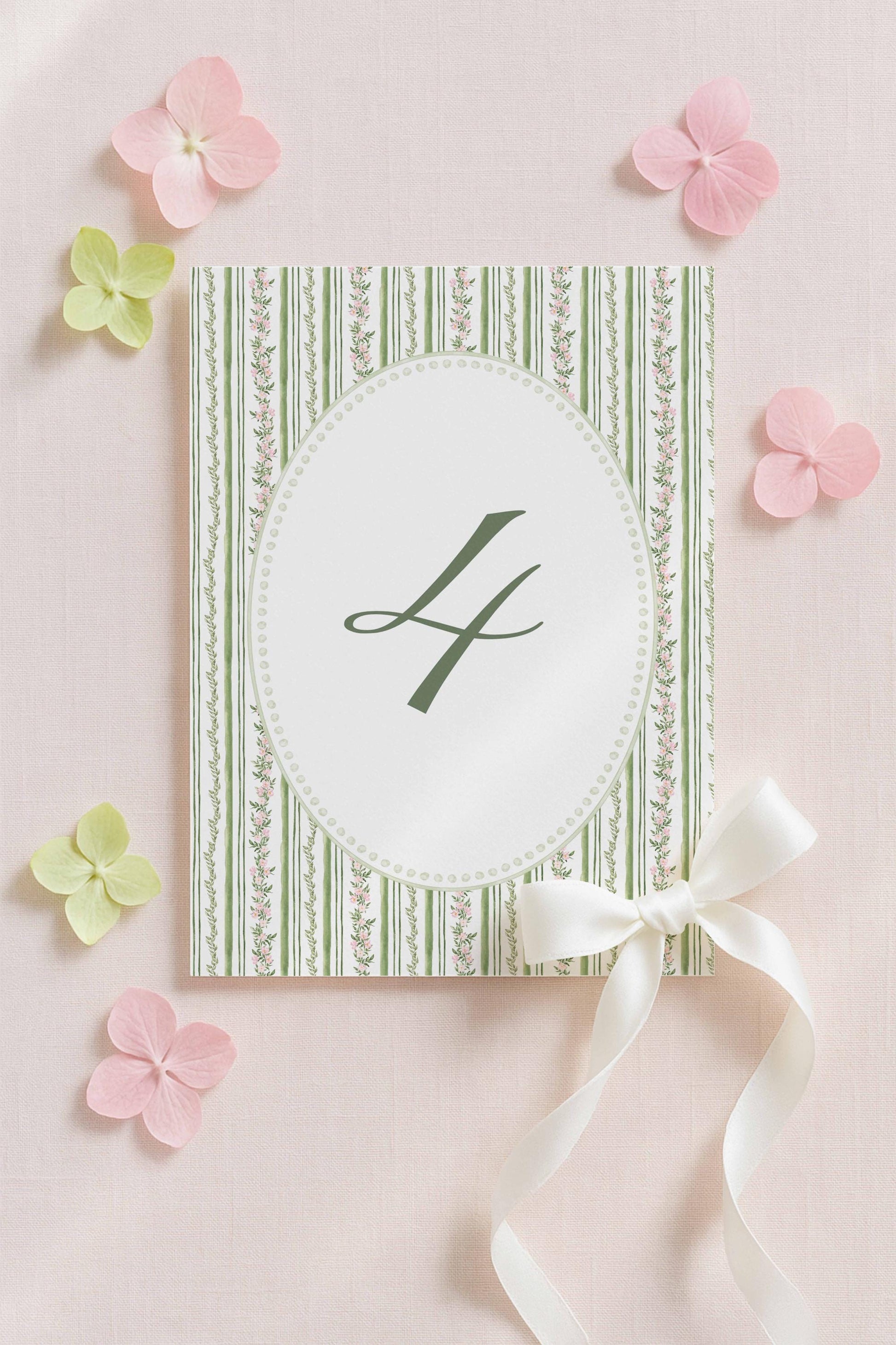 a white card with a green and white striped border, a green "L" in the center, and a white ribbon bow on the right side. The card is surrounded by pink flowers and other floral elements, creating a visually appealing and elegant composition.