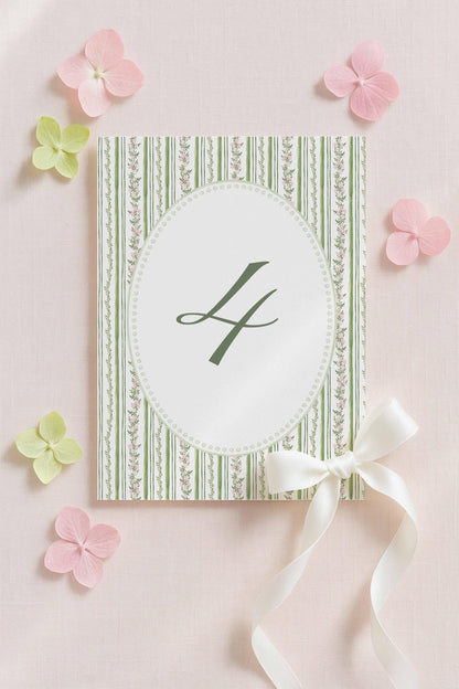 a white card with a green and white striped border, a green "L" in the center, and a white ribbon bow on the right side. The card is surrounded by pink flowers and other floral elements, creating a visually appealing and elegant composition.
