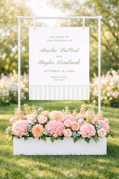 a white sign with text and a white planter filled with pink and white flowers in the foreground, set against a backdrop of trees and greenery.
