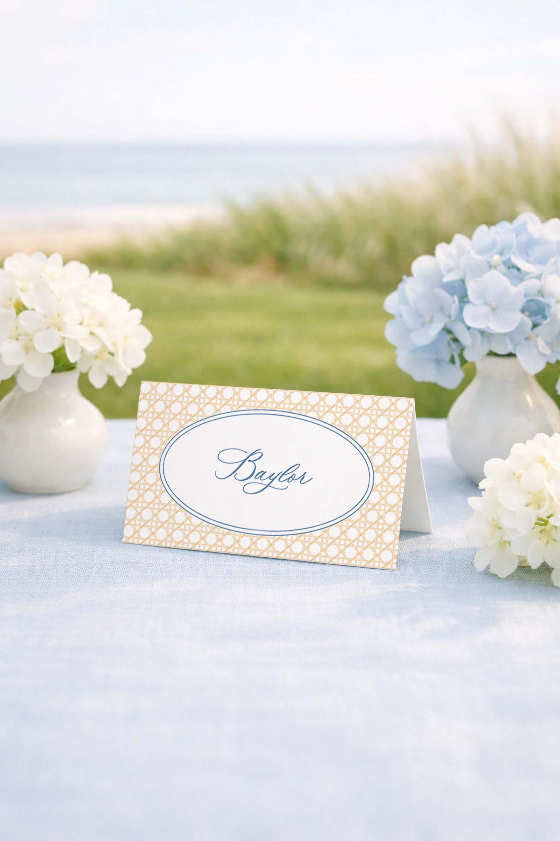 a table with a white tablecloth, a card with the name "Baylor" written on it, and two vases of white flowers. The background includes a grassy area and a body of water, creating a serene and picturesque setting.