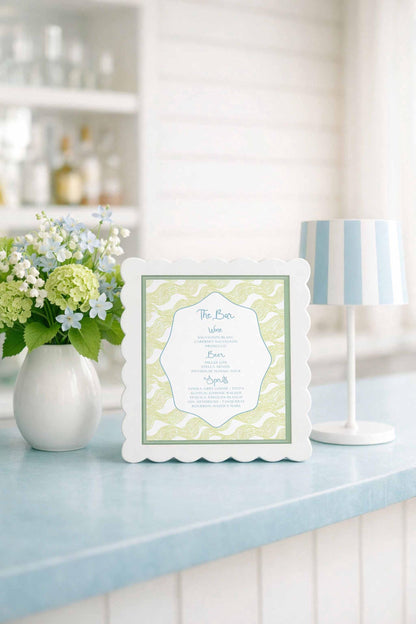 a white framed menu with a decorative border, placed on a blue countertop next to a vase of white flowers and a white lamp.