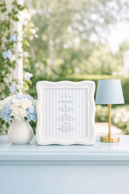 a white framed sign with blue stripes, placed on a light blue surface, next to a white vase filled with blue flowers and a small blue lamp.
