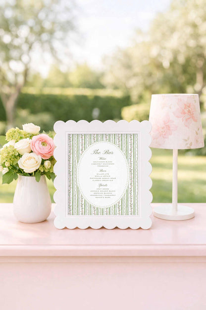 a white frame with a floral pattern, a vase of white and pink flowers, and a pink lamp on a pink table.