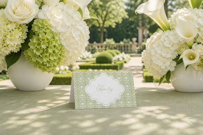 A table with a green card in the center, surrounded by two large vases filled with white flowers, including roses and hydrangeas, in a garden setting.